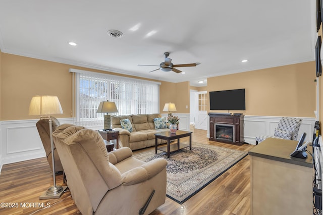 living room featuring crown molding, visible vents, a glass covered fireplace, wainscoting, and wood finished floors