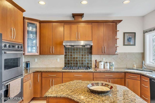 kitchen with under cabinet range hood, light stone counters, a sink, appliances with stainless steel finishes, and brown cabinetry