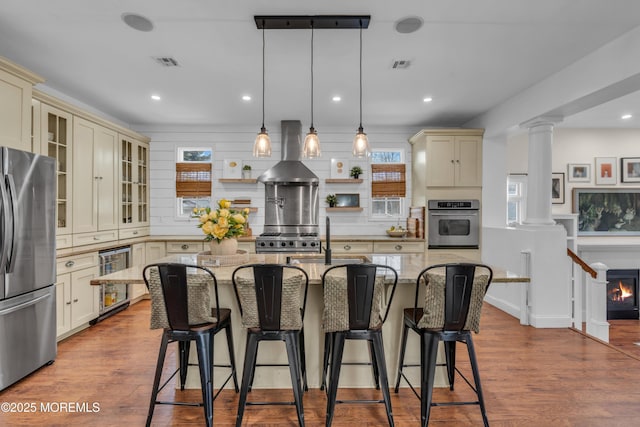 kitchen with wine cooler, cream cabinetry, stainless steel appliances, ornate columns, and wall chimney exhaust hood