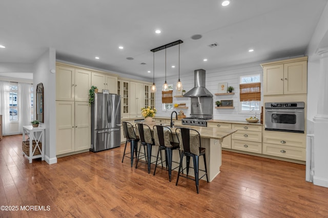 kitchen featuring light stone counters, wood-type flooring, cream cabinets, appliances with stainless steel finishes, and wall chimney range hood