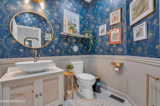 half bathroom featuring wallpapered walls, visible vents, wainscoting, toilet, and vanity