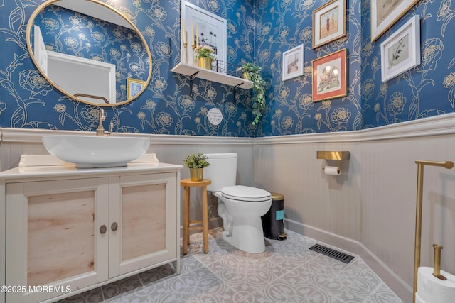 bathroom featuring visible vents, toilet, wainscoting, vanity, and wallpapered walls