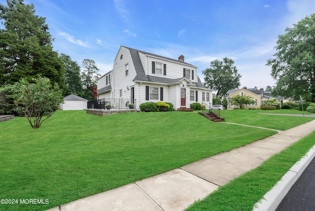 colonial inspired home featuring a shingled roof, an outdoor structure, a gambrel roof, a chimney, and a front yard