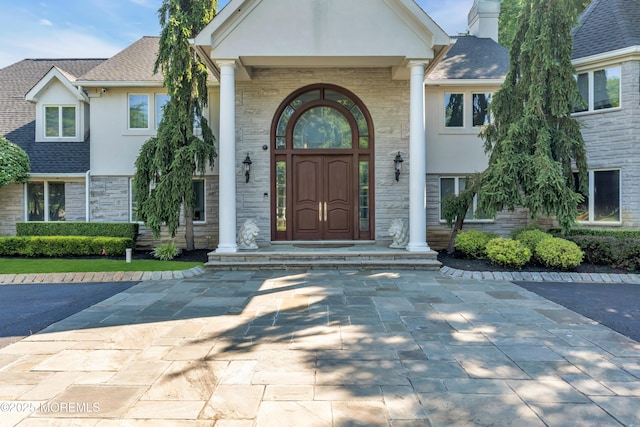 view of exterior entry with stone siding, roof with shingles, and stucco siding