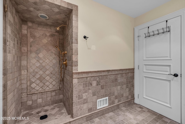 bathroom featuring a tile shower, visible vents, wainscoting, and tile walls