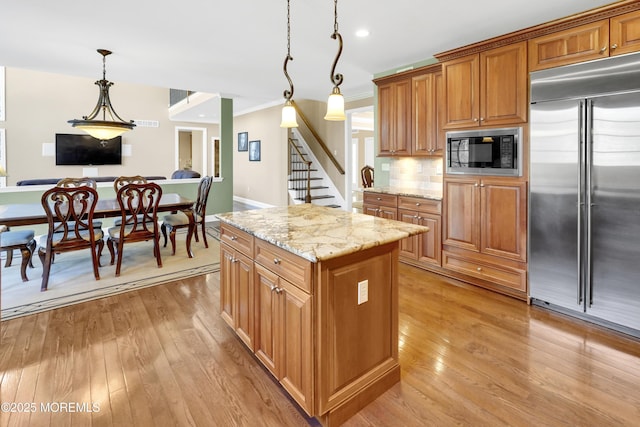 kitchen with light wood finished floors, hanging light fixtures, brown cabinetry, light stone countertops, and built in appliances