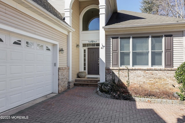 doorway to property featuring an attached garage, brick siding, decorative driveway, and a shingled roof