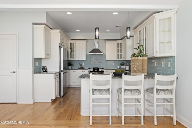 kitchen with a breakfast bar area, appliances with stainless steel finishes, white cabinetry, a peninsula, and wall chimney exhaust hood