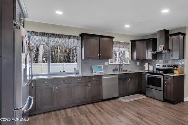 kitchen featuring appliances with stainless steel finishes, a sink, wall chimney range hood, dark brown cabinets, and wood finished floors