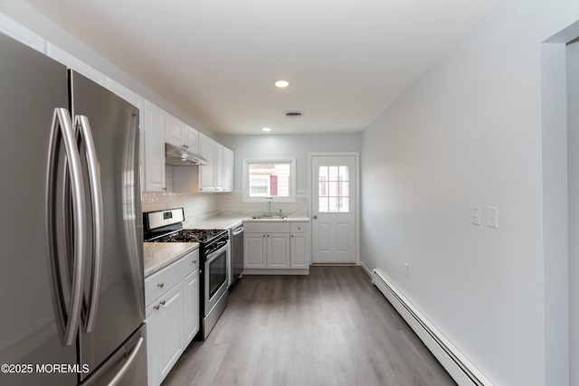 kitchen with a baseboard radiator, under cabinet range hood, stainless steel appliances, a sink, and decorative backsplash