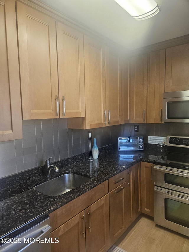 kitchen with stainless steel appliances, a sink, light wood-style floors, decorative backsplash, and dark stone counters