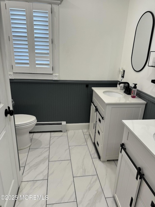 bathroom with two vanities, a sink, marble finish floor, baseboard heating, and wainscoting