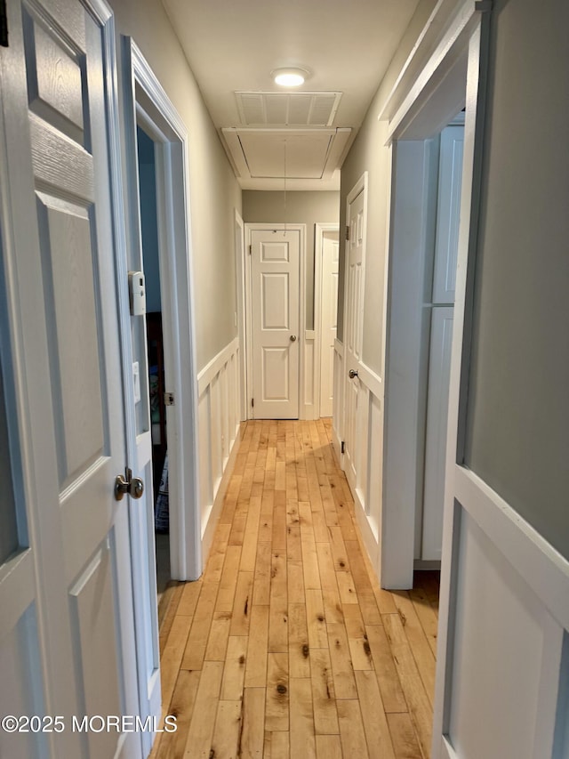 hallway with light wood-style flooring, a decorative wall, visible vents, wainscoting, and attic access