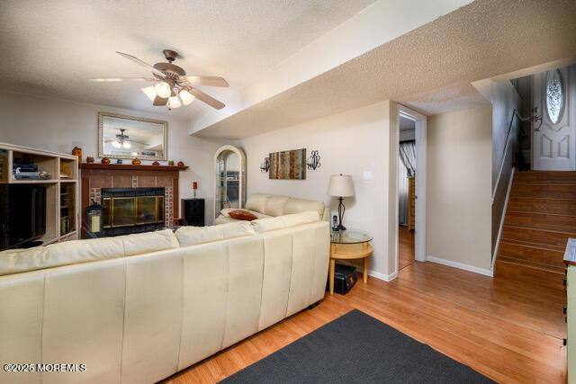 living area featuring a ceiling fan, a brick fireplace, a textured ceiling, wood finished floors, and baseboards