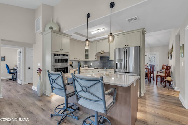 kitchen with light wood-style flooring, under cabinet range hood, appliances with stainless steel finishes, backsplash, and a raised ceiling