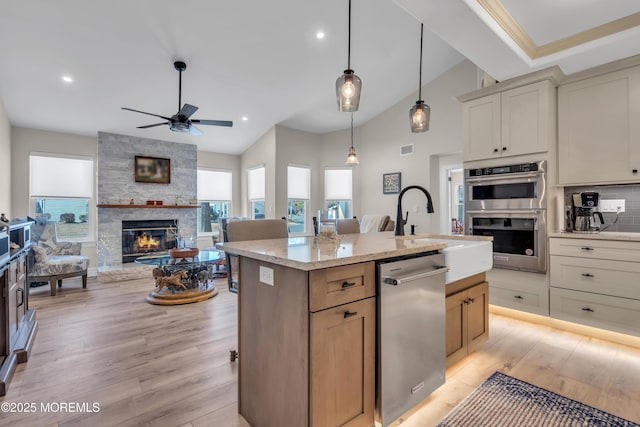 kitchen featuring decorative light fixtures, stainless steel appliances, light wood-style floors, a kitchen island with sink, and a stone fireplace