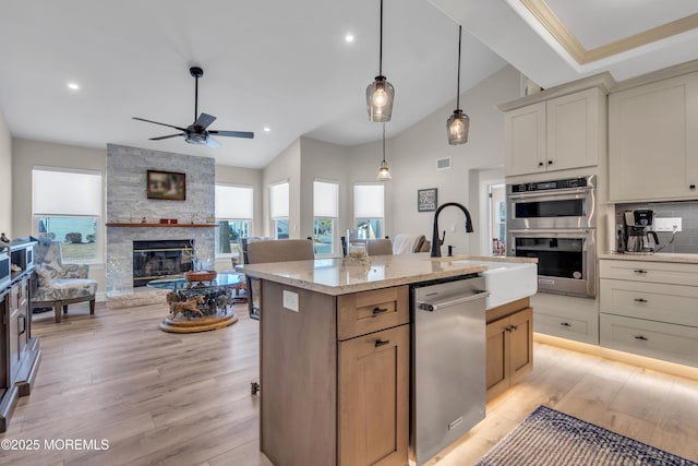 kitchen featuring light wood finished floors, decorative backsplash, an island with sink, appliances with stainless steel finishes, and a stone fireplace