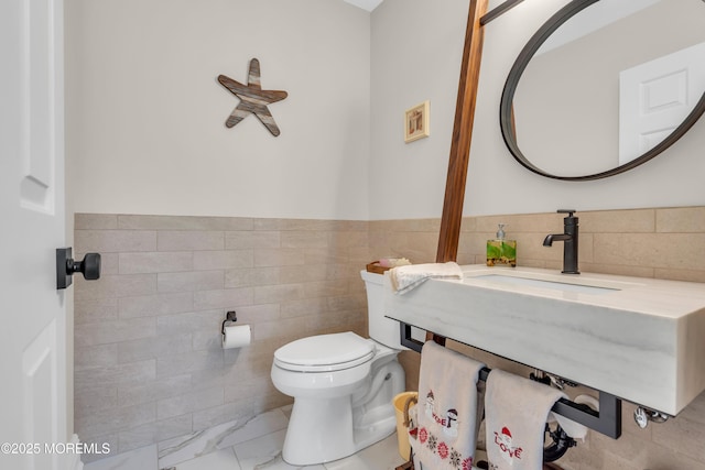 half bathroom with wainscoting, toilet, a sink, marble finish floor, and tile walls