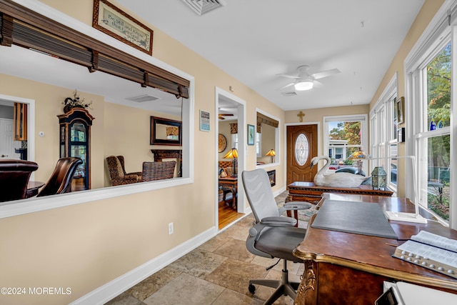 office featuring ceiling fan, stone tile floors, visible vents, baseboards, and a glass covered fireplace