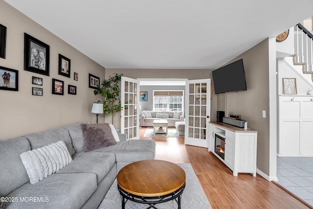 living room featuring baseboards, a glass covered fireplace, stairway, french doors, and light wood-style floors