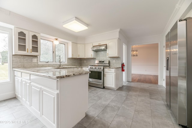 kitchen featuring light stone countertops, under cabinet range hood, a peninsula, appliances with stainless steel finishes, and decorative backsplash