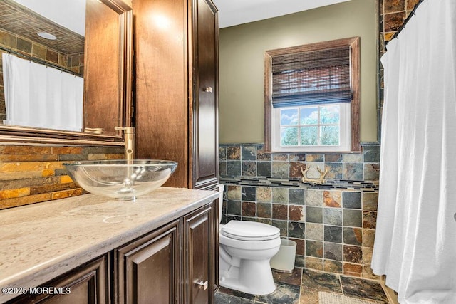 bathroom featuring wainscoting, toilet, stone finish floor, vanity, and tile walls