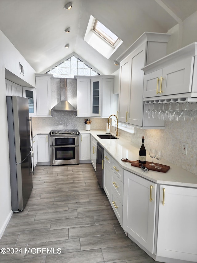 kitchen featuring vaulted ceiling with skylight, stainless steel appliances, a sink, wall chimney range hood, and decorative backsplash