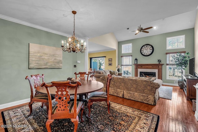 dining room featuring ceiling fan with notable chandelier, hardwood / wood-style flooring, a warm lit fireplace, crown molding, and baseboards