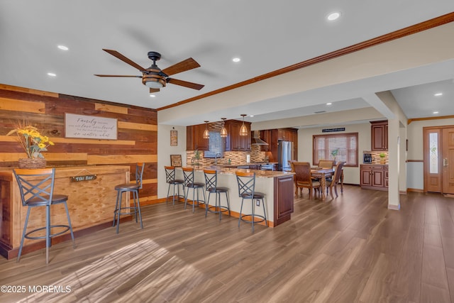 kitchen with wood finished floors, a peninsula, freestanding refrigerator, a kitchen breakfast bar, and wall chimney exhaust hood