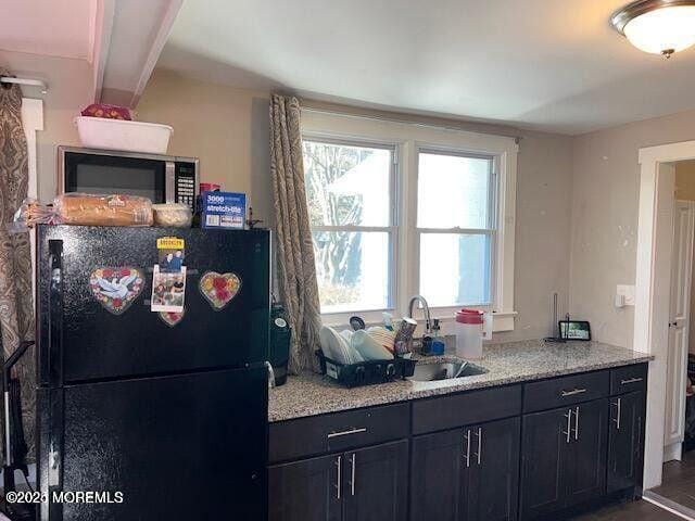 kitchen with stainless steel microwave, dark cabinets, freestanding refrigerator, light stone countertops, and a sink