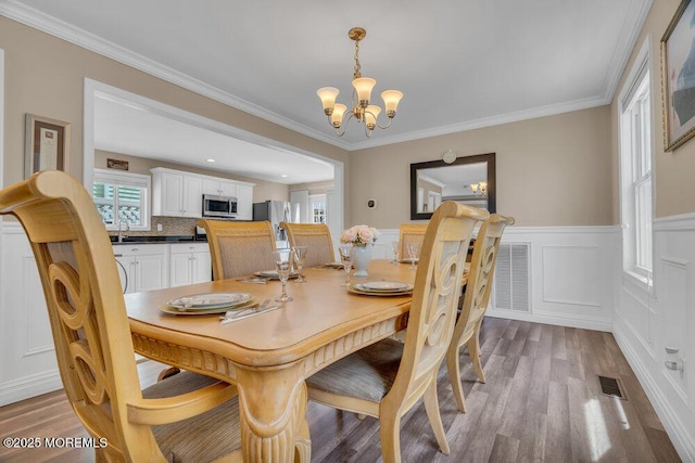 dining room featuring a chandelier, a wainscoted wall, wood finished floors, visible vents, and crown molding