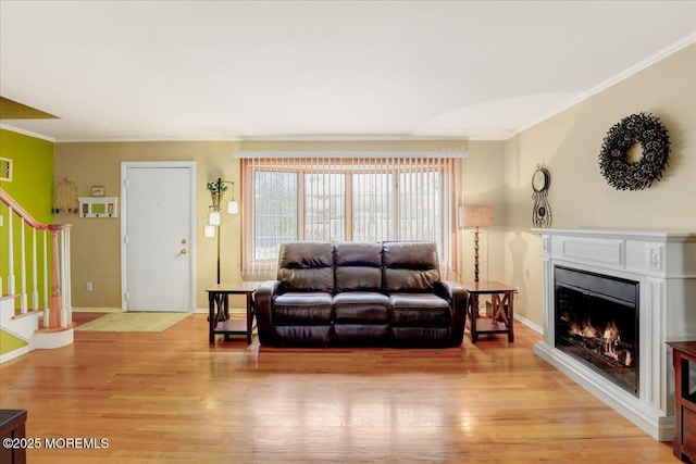 living room with a warm lit fireplace, baseboards, light wood-style flooring, stairs, and crown molding