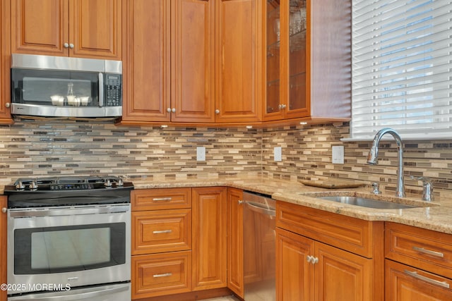 kitchen featuring backsplash, glass insert cabinets, light stone counters, appliances with stainless steel finishes, and a sink