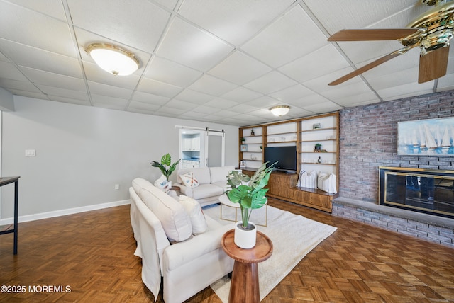 living room with built in features, a fireplace, a barn door, a drop ceiling, and baseboards