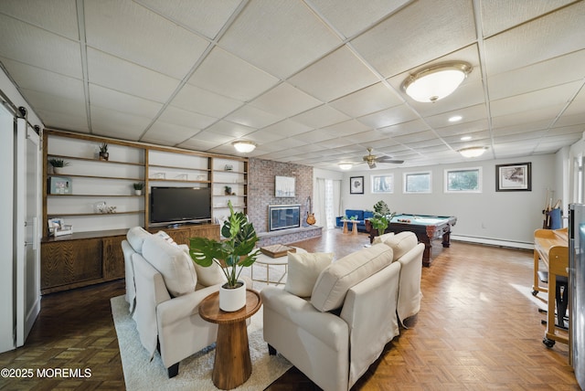 living room featuring built in shelves, pool table, baseboard heating, a brick fireplace, and a drop ceiling