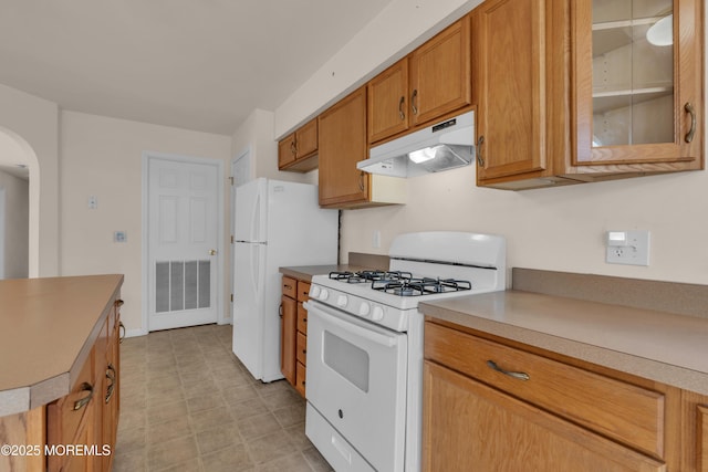 kitchen with white appliances, brown cabinetry, arched walkways, glass insert cabinets, and under cabinet range hood