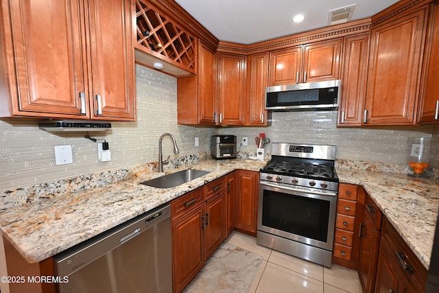 kitchen featuring brown cabinetry, light stone counters, a sink, stainless steel appliances, and backsplash