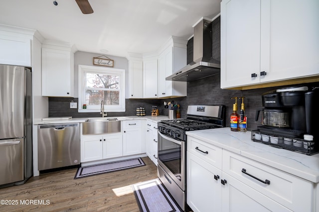 kitchen with wood finished floors, a sink, stainless steel appliances, white cabinetry, and wall chimney exhaust hood