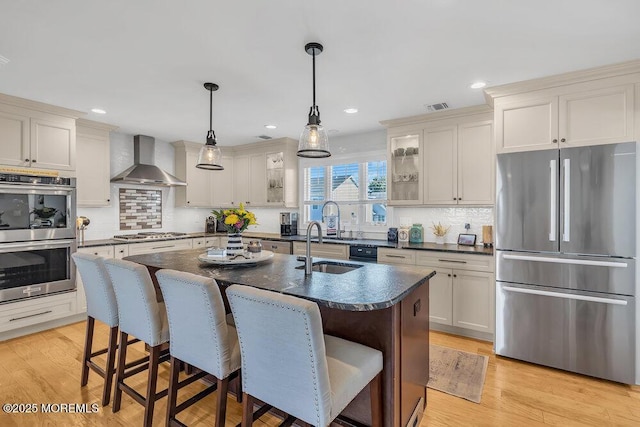 kitchen with light wood finished floors, an island with sink, stainless steel appliances, a kitchen breakfast bar, and wall chimney exhaust hood