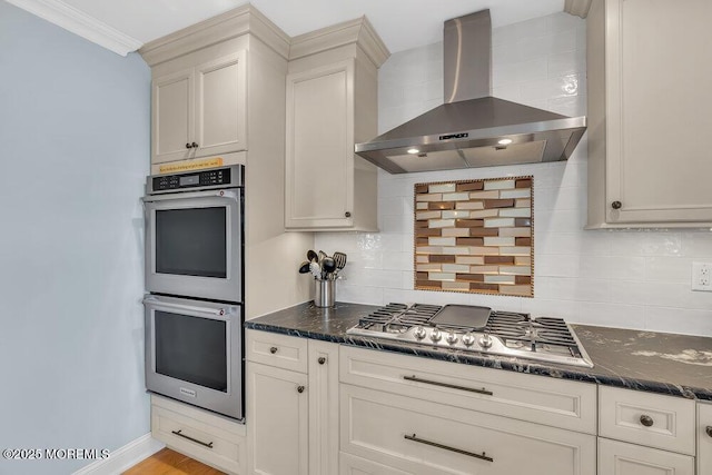 kitchen featuring backsplash, crown molding, wall chimney range hood, dark stone countertops, and appliances with stainless steel finishes