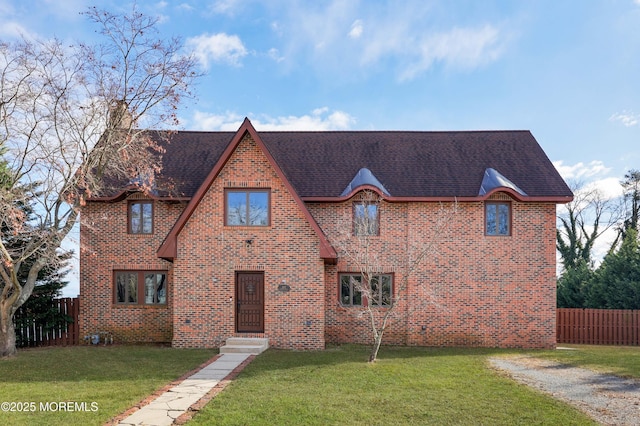 tudor-style house featuring brick siding, fence, roof with shingles, and a front yard