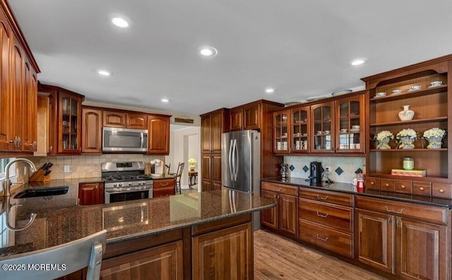 kitchen with a sink, dark stone countertops, light wood-style floors, appliances with stainless steel finishes, and glass insert cabinets