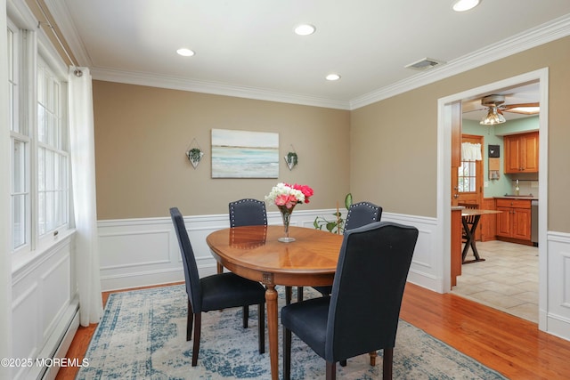 dining room with light wood finished floors, visible vents, crown molding, a baseboard heating unit, and a wainscoted wall