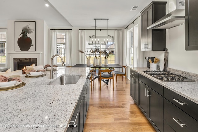 kitchen featuring visible vents, stainless steel gas cooktop, a lit fireplace, wall chimney exhaust hood, and a sink