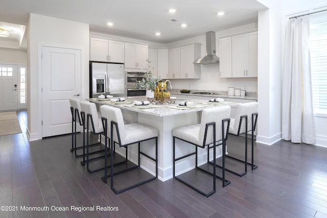 kitchen with a kitchen bar, a center island, appliances with stainless steel finishes, wall chimney exhaust hood, and dark wood-style flooring