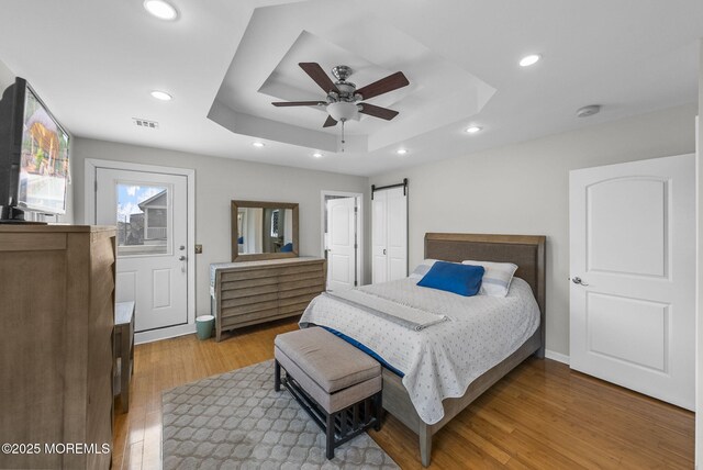 bedroom with wood finished floors, recessed lighting, a barn door, a raised ceiling, and ceiling fan