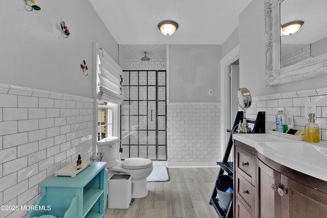 bathroom with a wainscoted wall, toilet, vanity, tiled shower, and tile walls