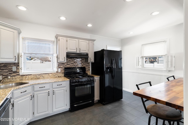 kitchen featuring black appliances, a sink, light stone counters, under cabinet range hood, and wainscoting