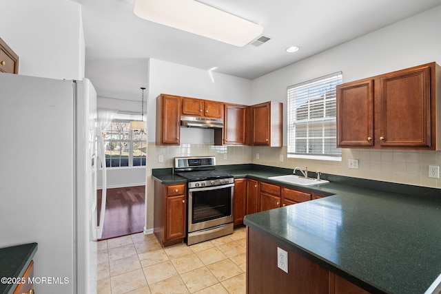 kitchen with under cabinet range hood, a sink, dark countertops, freestanding refrigerator, and stainless steel range with gas cooktop