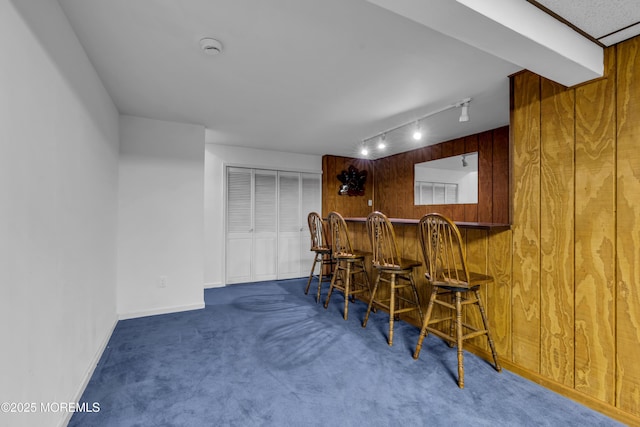 bar featuring baseboards, a bar, rail lighting, wood walls, and carpet flooring